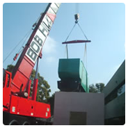 A red crane lifting a green metal ups power supply near a building
